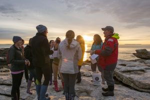 Students and the instructor gather together on the rocky shore at sunset.