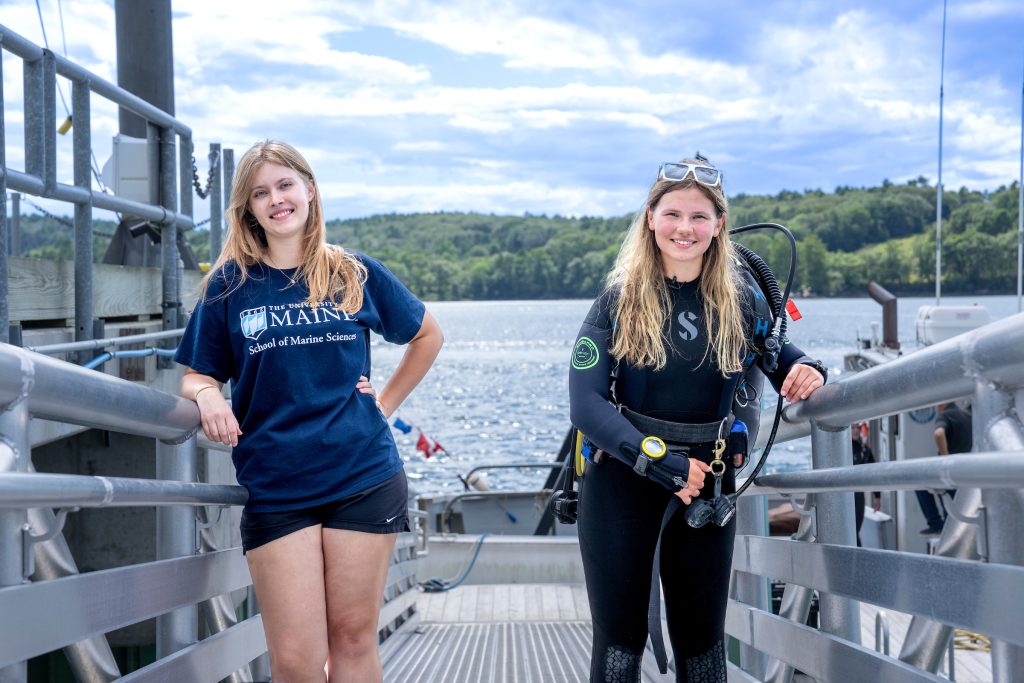 Two UMaine students, one in a Marine Science shirt and the other in scuba gear standing at the dock.