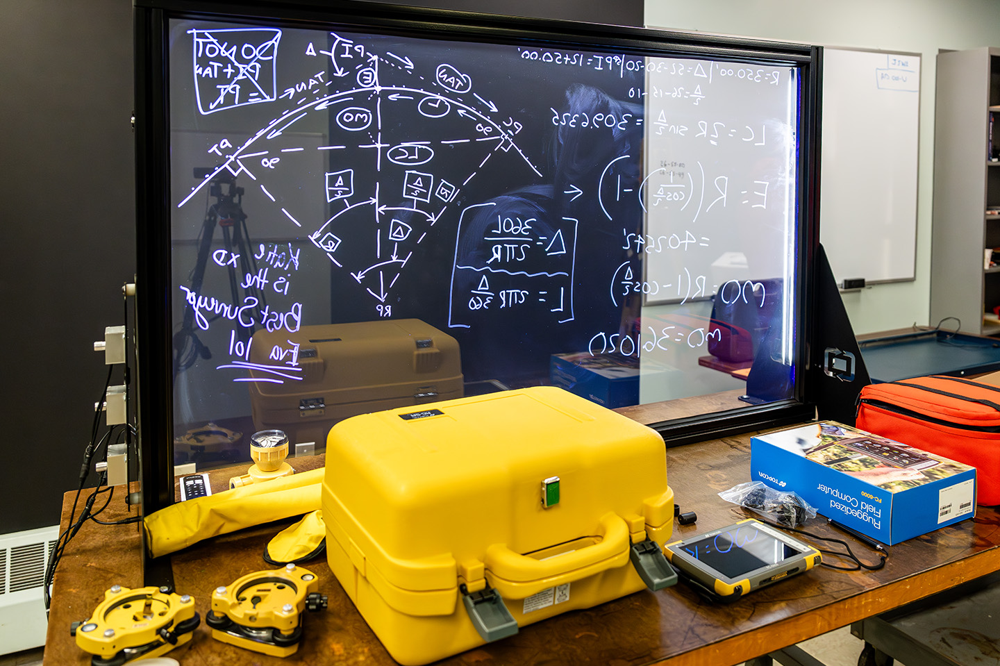 Surveying Engineering Technology materials on a table with written out math equations on a board