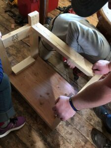 Now the team is making sure that the leg supports are in place to keep the table strong and durable. Some members are holding the table in place while another is screwing the supports in place.