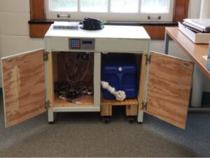 The cabinet doors of the system are opened so the bottom shelves are visible. The water tank os located on the right of the shelf, and the pump, manifolds, and solenoids are located to the left.