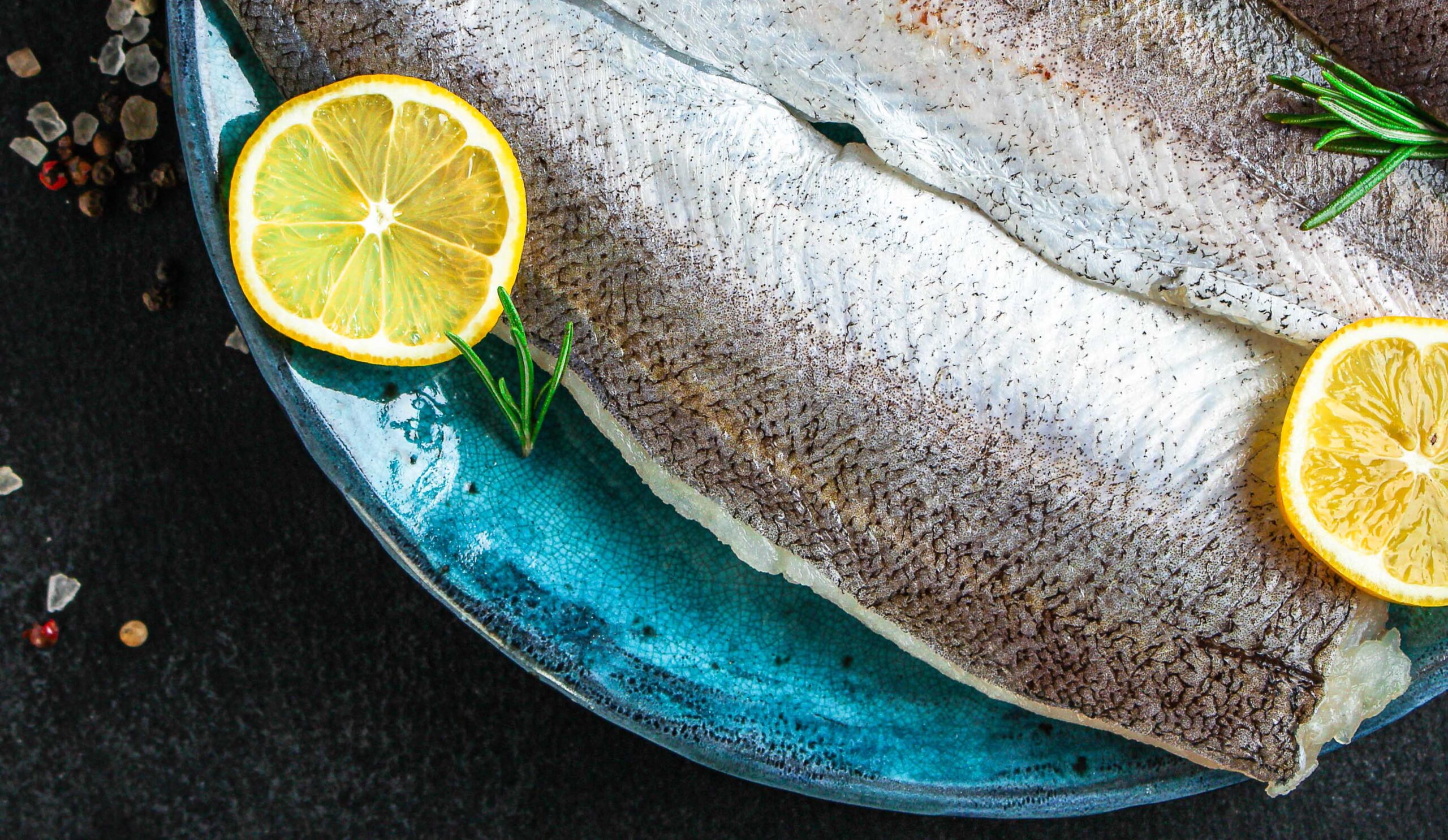 Raw hake fish fillet on turquoise plate with lemon slices on black background