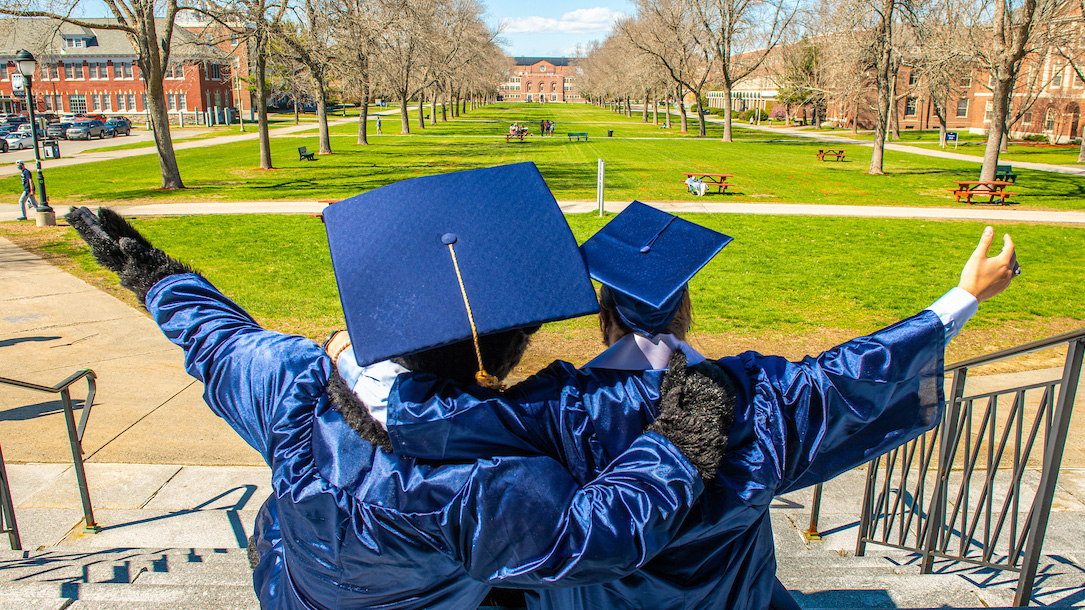 A student poses with the UMaine mascot Bananas, both dressed in graduation cap and gown, overlooking the UMaine campus mall from the steps of Fogler Library