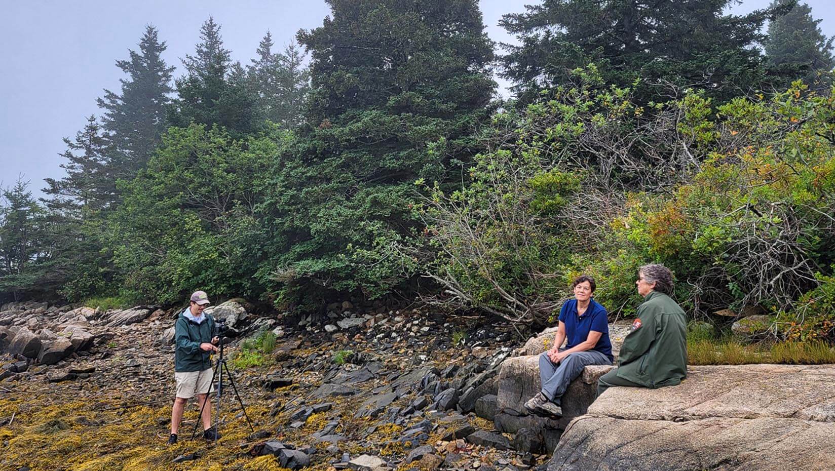 A photo of two people being recorded on video while sitting on a rock on the Maine coast