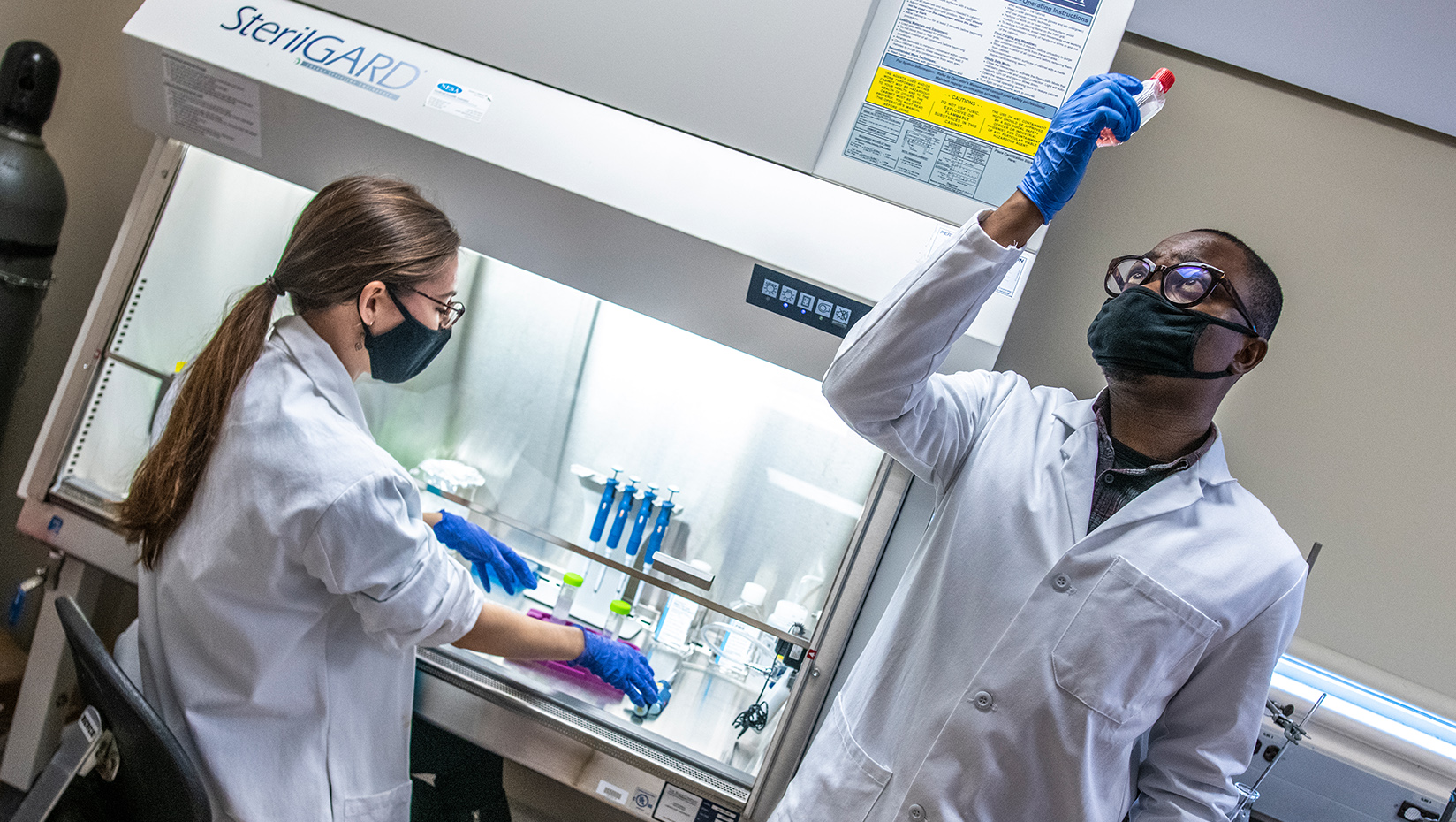 Two lab partners in white coats, gloves, glasses, and masks are in the midst of testing liquids in viles. One holds a bottle of liquid up to the light, and one is sitting down in front of a lab station.