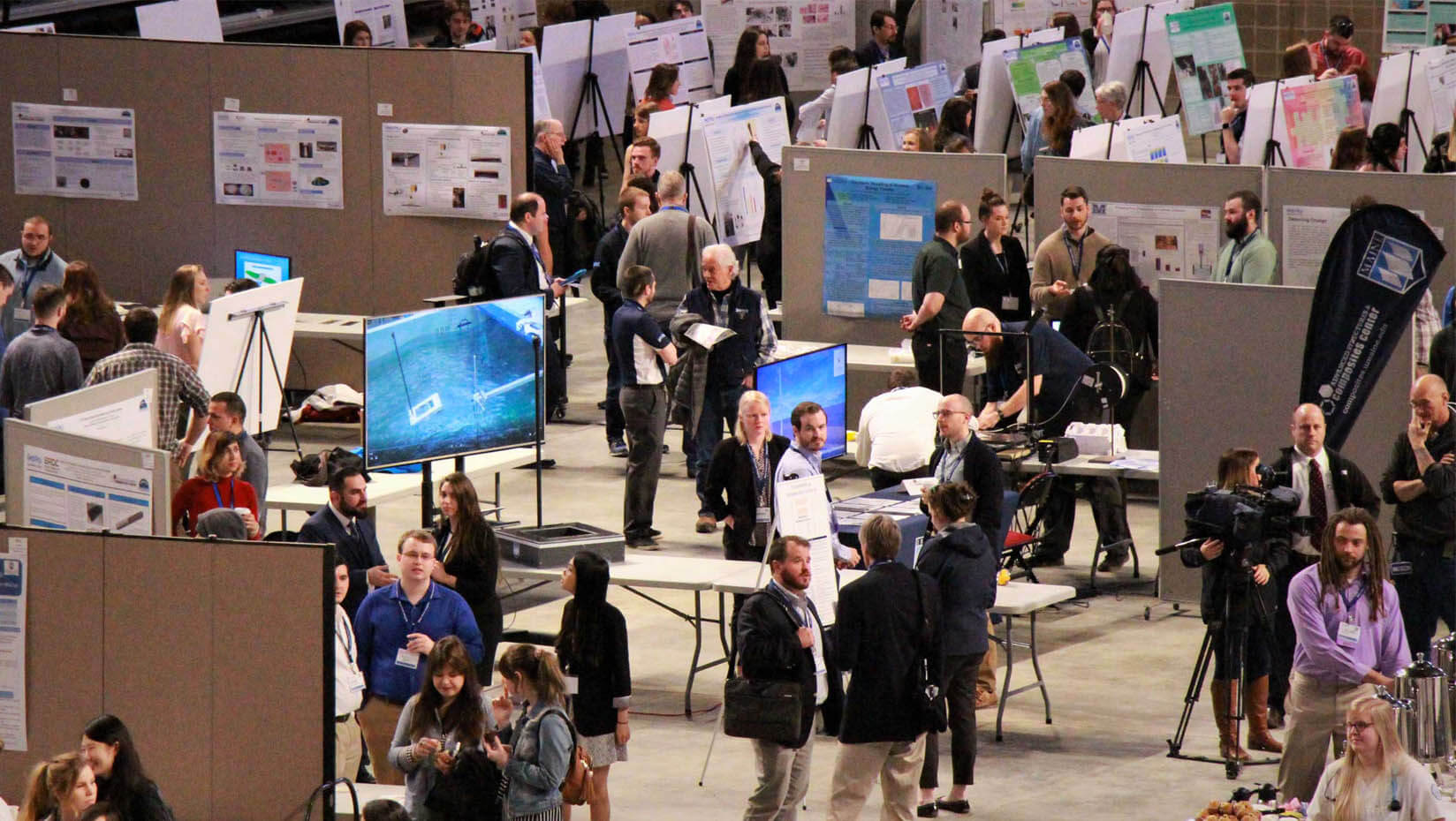 A photo of presenters and attendees standing in a large room with posters