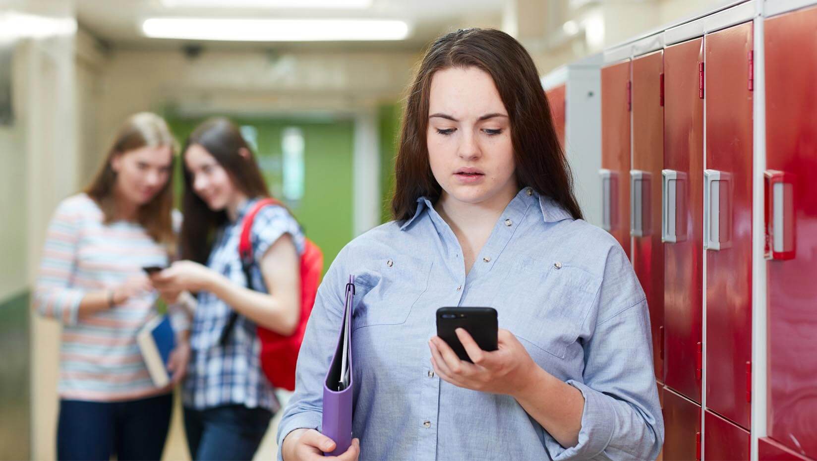 A photo showing two people cyberbullying a third person in a school hallway lined with lockers