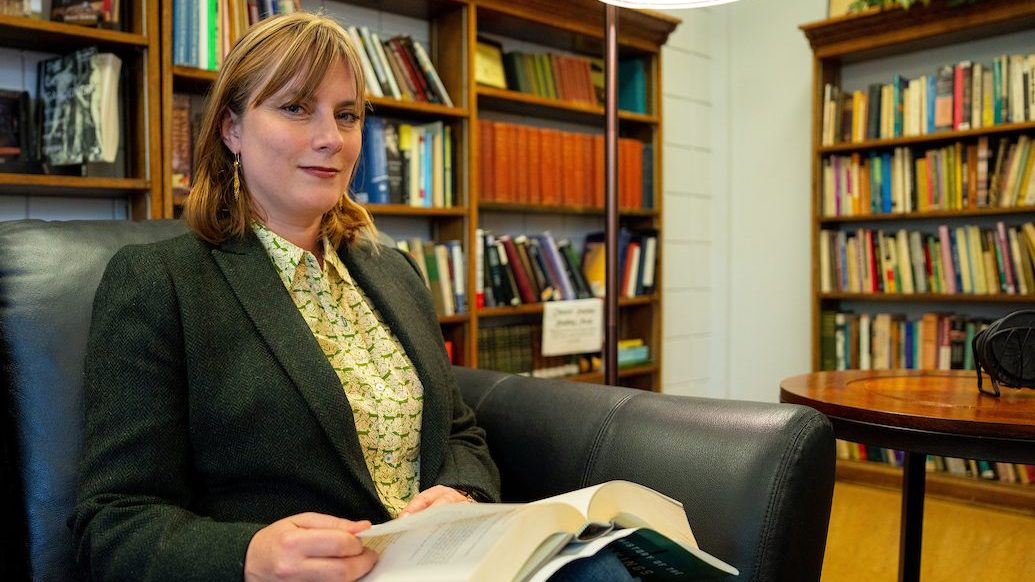 A photo Sarah Harlan-Haughey, seated in a library with a book on her lap