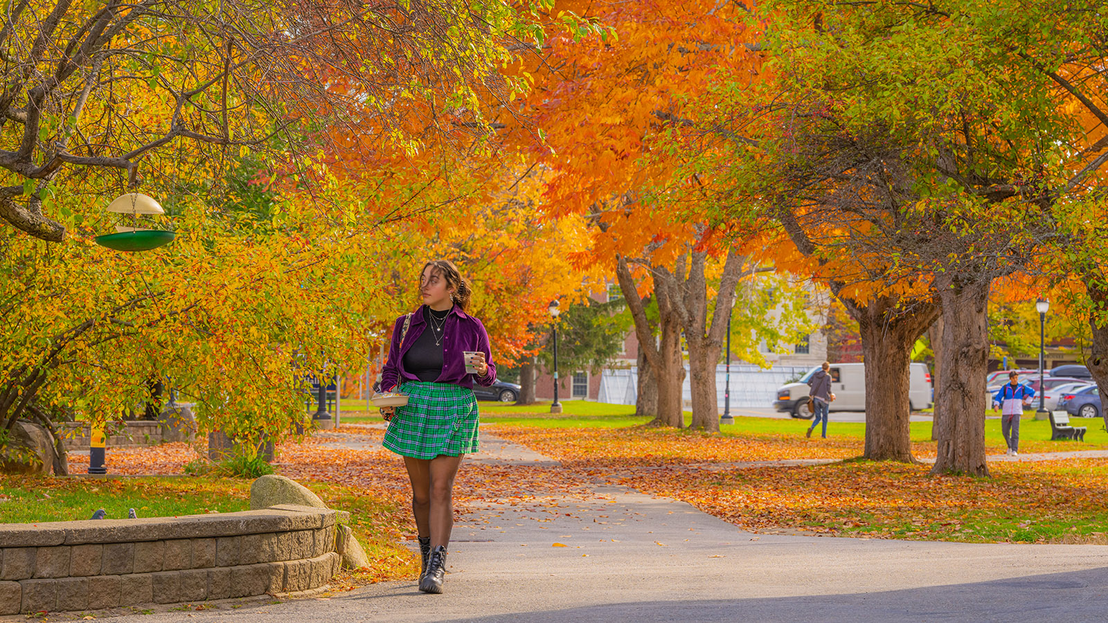 An outdoor photo of the UMaine campus taken October 2022, depicting a student walking a path on campus among the fall foliage