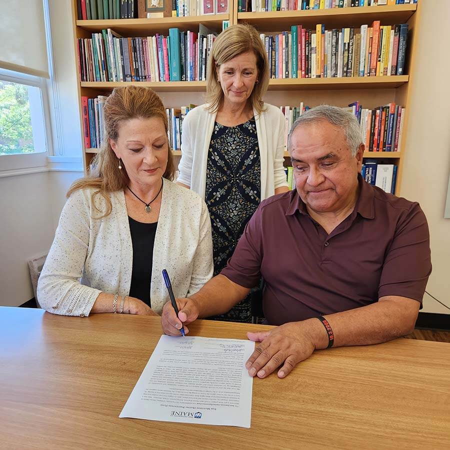 A photo of Jolayne Houtz and Hector Martinez signing a document while Elizabeth Allen looks on.