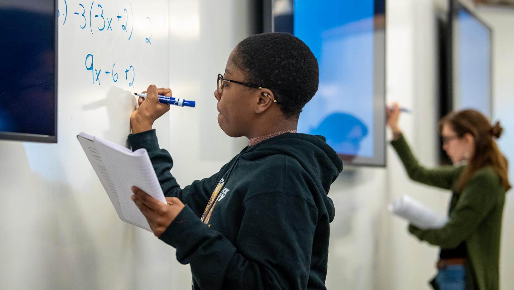 A photo of two students writing math equations on a white board