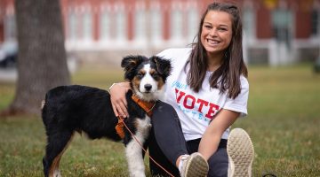 A photo of a person sitting with a dog outside on the University of Maine Mall.