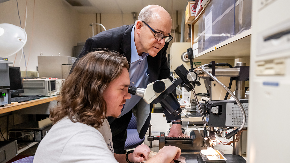 A professor looks on as a student sits at a magnification scope in an electrical and computer engineering lab, examining a circuit board.