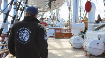 A photo of a person wearing a UMaine Aquaculture Research Institute jacket on the deck of a tallship