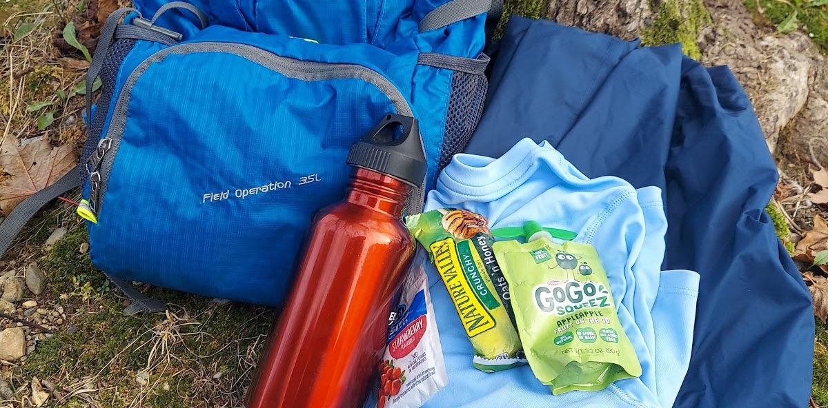 Photo of a backpack leaning up on a tree. On the ground beside the bag is a water bottle, a change of clothes and some snacks.