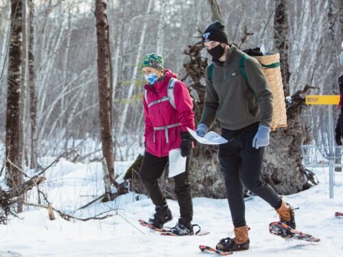 Two students snowshoe in the woods while holding maps