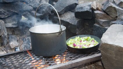 A pot and fry pan of cooking food on a campfire with a grate and rocks