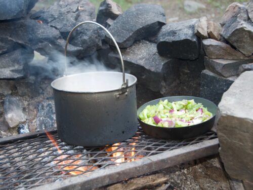 A pot and fry pan of cooking food on a campfire with a grate and rocks