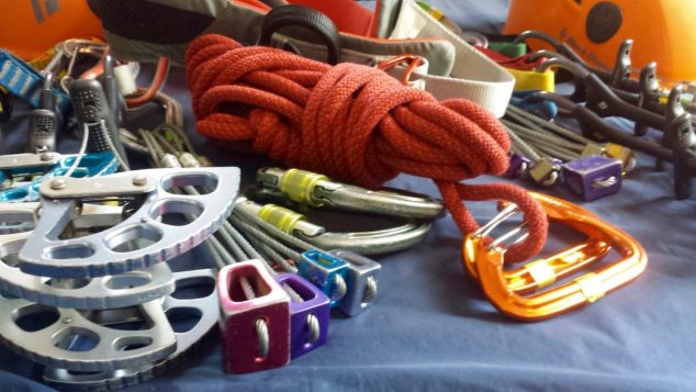 Image of climbing gear: (from left to right) a Cam, some nuts, carabiners, accessory cord, a harness, more nuts, more cams and a helmet.
