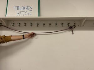 Rope hanging off of hangers below a sign that says 'trucker's hitch' with a hand holding the rope