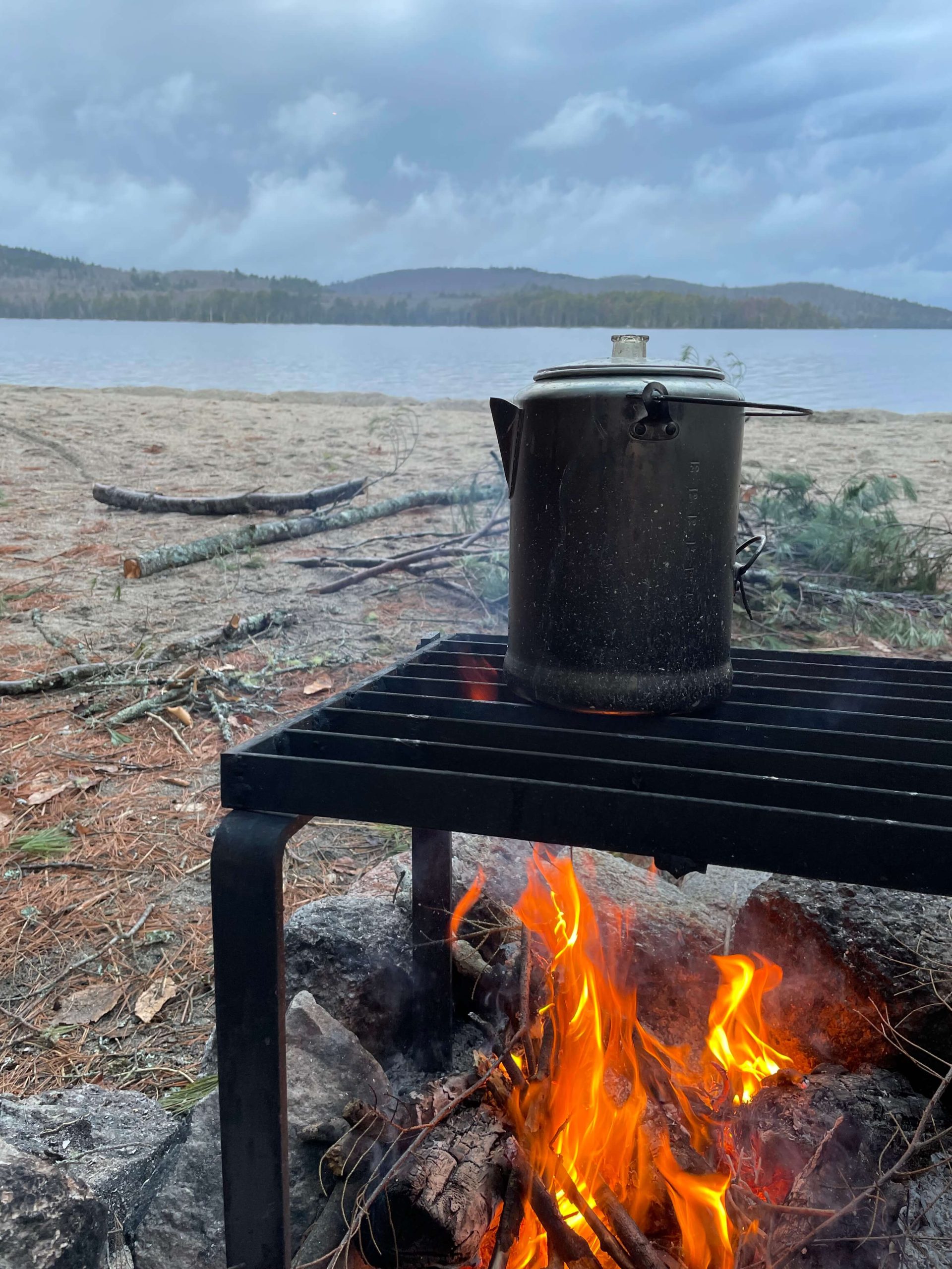 A kettle of hot water is cooking over an open fire on a campsite located on a beach next to a lake.