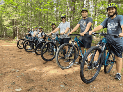 A row of students on mountain bikes. They are lined up side by side on a dirt trail in the woods during the day.