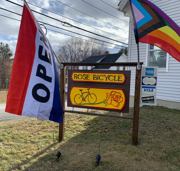 A sign for Rose Bike Shop with a flag that reads "Open" and an LGBTQIA+ flag waiving on either side of the sign