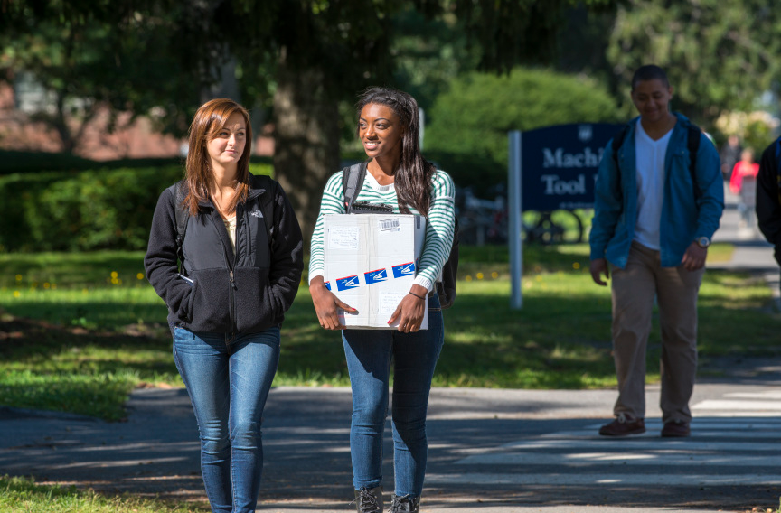 Two students walk on a campus path; one holds a USPS package.