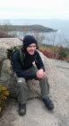 A Young Adult man smiling while sitting in hiking gear on the summit of a hill or mountain. With a scenic overlook in the background of a coast.