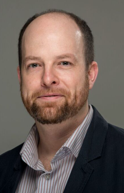 A man (Don Beith) in a collared shirt and blazer, smiling. With a grey background.