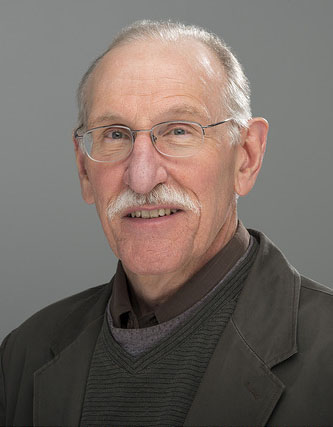 Older mustached man ( Michael Howard) wearing glasses and a blazer.