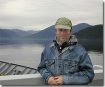 Adult man (Roger King) in a jean jacket in a hat with a slight smile. In the background, there is a lake/pond and a range of mountains/hills.