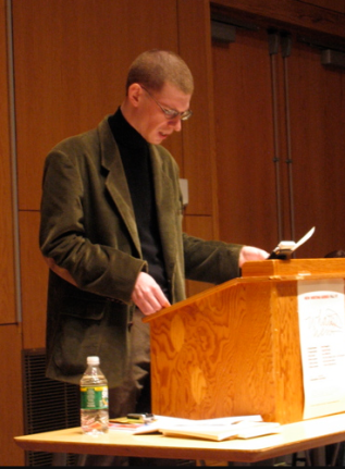 Aaron Kunin reading aloud behind a podium