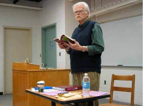 Robert Kelly reading aloud from a book in hand while standing behind a desk