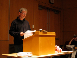 Rod Smith reading off a piece of paper aloud to an audience behind a podium