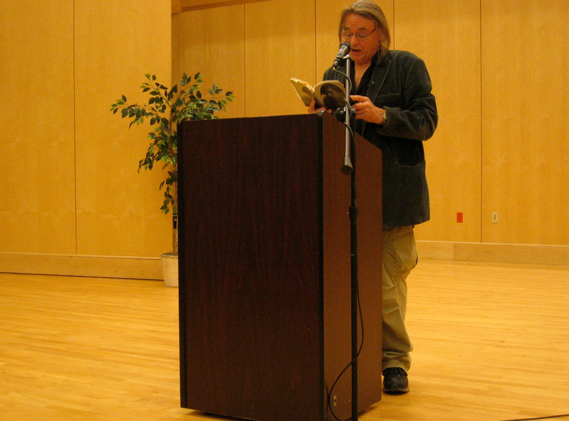 Tom Pickard reading out of a book into a microphone behind a podium