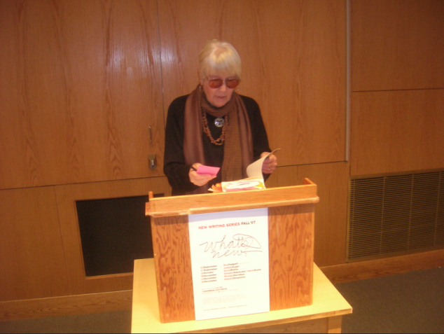 Joanne Kyger reading out of her book behind a podium