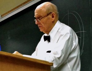 Professor John Nickerson standing at podium in white shirt with bow tie.