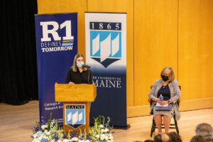 Valeria Roach at the podium of the 2022 State of the University Address while President Joan Ferrini-Mundy (seated at right) looks on.