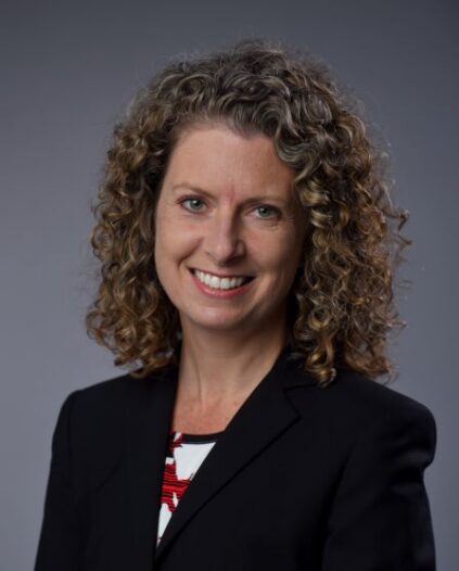 Professional headshot of Dean Penny Bishop smiling at camera, wearing a dark blazer, against a dark background.