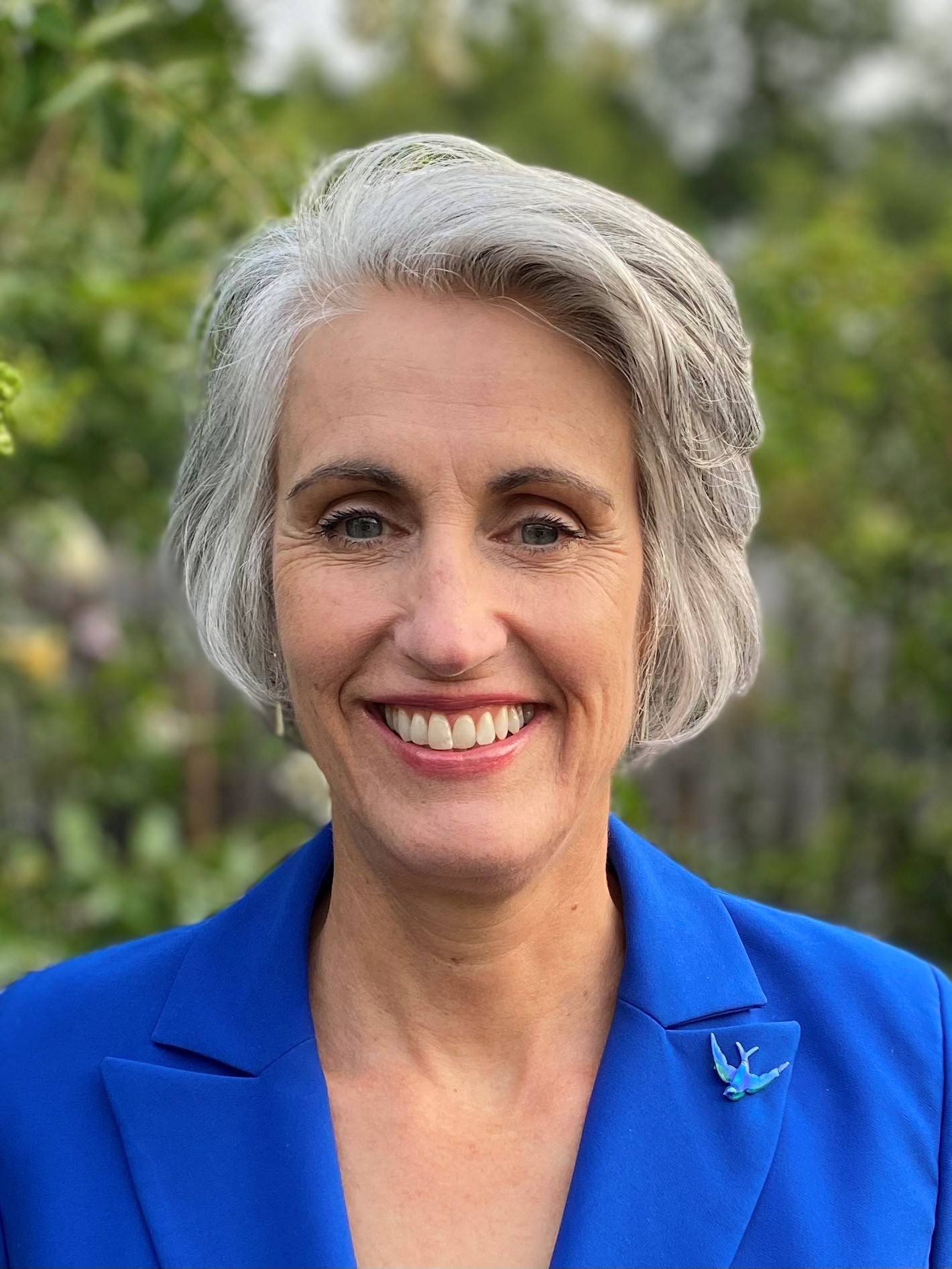 Professional headshot of Dean Diane Rowland, smiling at camera and wearing a blue jacket, in front of a leafy forest background.