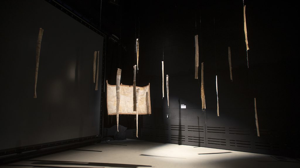 A photo of an art installation featuring beige fabric scraps hanging from the ceiling of a black room. A beam of light illuminates some of the fabric.