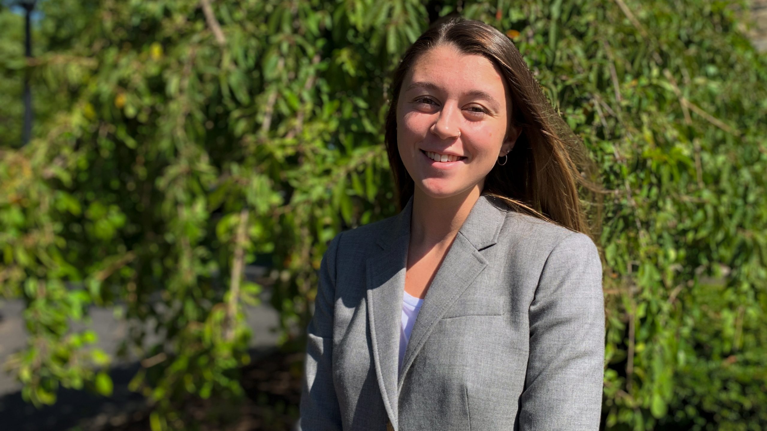 Lola Holcomb poses in grey jacket in front of greenery