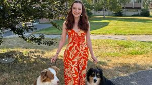 Amanda Ignacz stands in a red and gold summer dress with two dogs sitting on either side of her