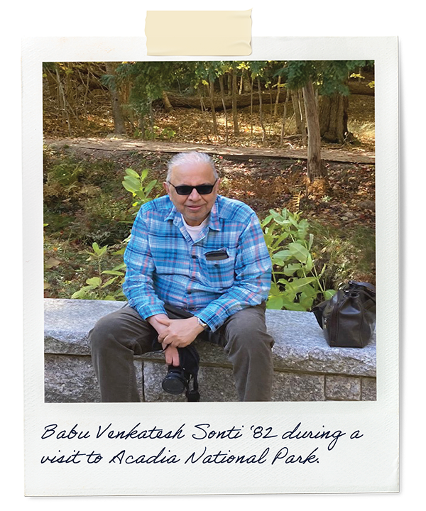 Babu sits on a trees bridge wall in front of trees. The caption says, "Babu Venkatesh Sonti ‘82 during a visit to Acadia National Park."