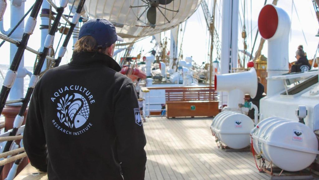 A photo of a person wearing a UMaine Aquaculture Research Institute jacket on the deck of a tallship