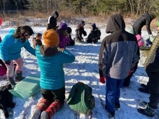 Students are dressed in outdoor weather gear and are sitting in a circle, engaged in an outdoor learning activity