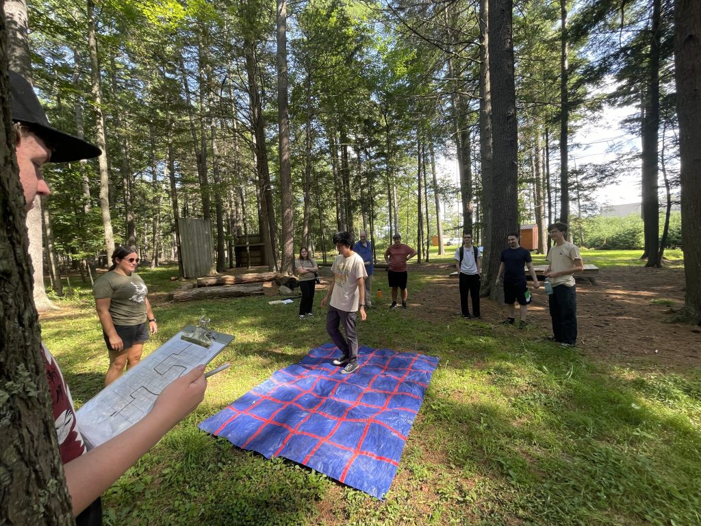 CCG - Maze Challenge & Cohort Building - students working together to navigate a grid outline on a tarp that is on the ground to solve a path drawn by another student.