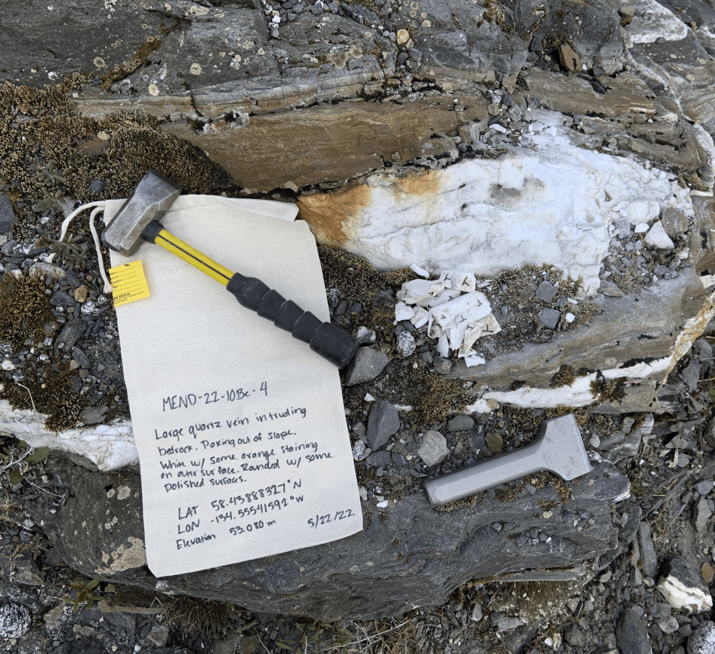 Scientific notebook and rock hammer on top of rock outcrop, showing a quartz vein.