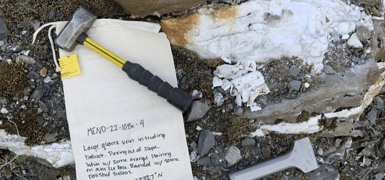 Scientific notebook and rock hammer on top of rock outcrop, showing a quartz vein.
